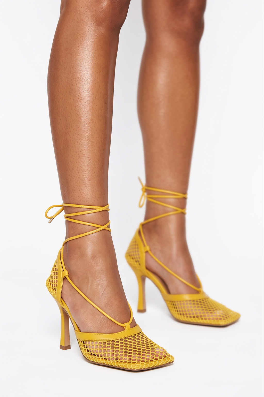 A side-by-side comparison of a model's legs wearing yellow openwork mesh high-heeled sandals with wraparound ankle straps, shown from the front and side angles against a plain white background.